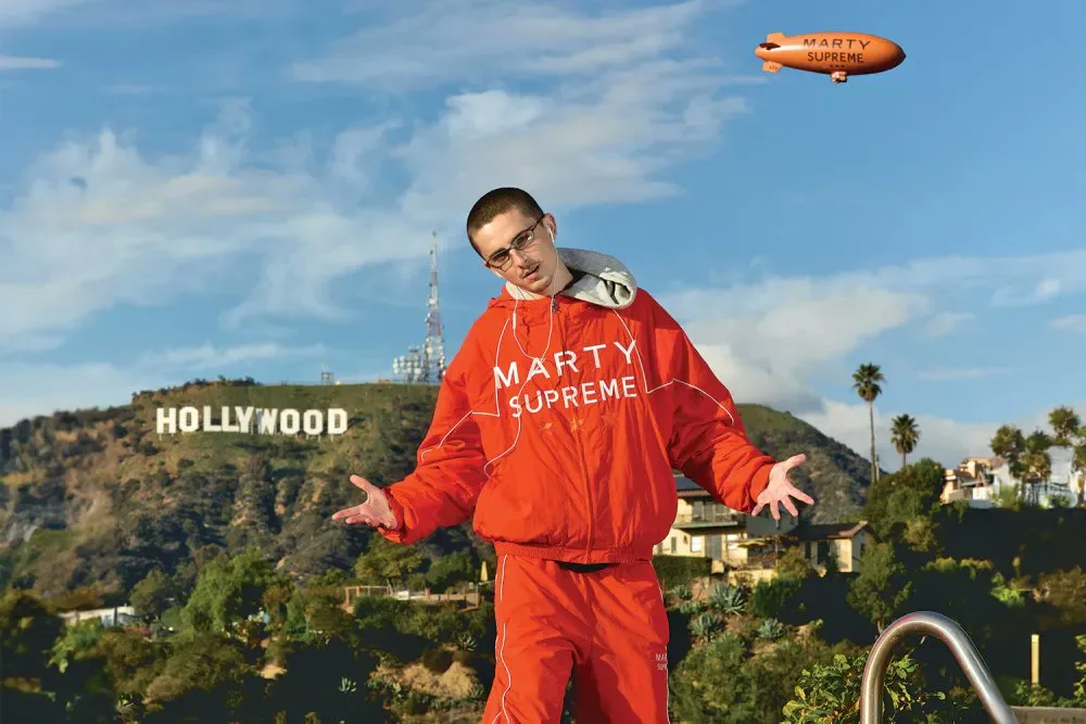 Timothée Chalamet posa con un chándal rojo de Marty Supreme frente al letrero de Hollywood. En el cielo azul destaca un dirigible naranja con el nombre de la película.