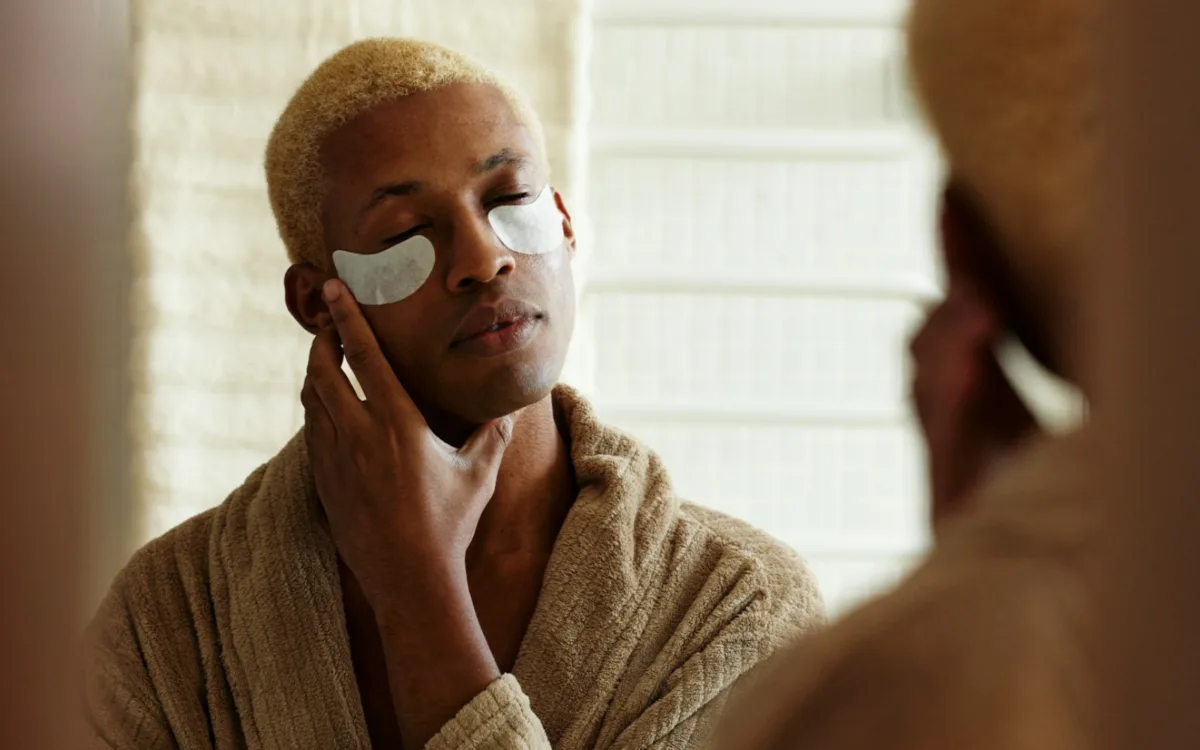 Hombre joven frente al espejo usando una bata de baño y parches hidratantes bajo los ojos como parte de su rutina de skincare masculino y cuidado facial para caballeros.