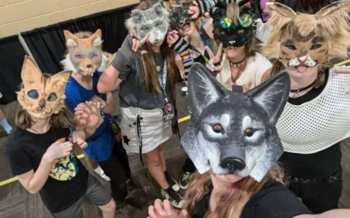 Siete jóvenes therians posan para una selfie en un salón, liderados por una therian que lleva una máscara de lobo gris y hace un gesto de garra. El grupo luce una gran variedad de máscaras de zorros y felinos, complementadas con ropa casual, pulseras coloridas y accesorios temáticos como una pata de felpa.