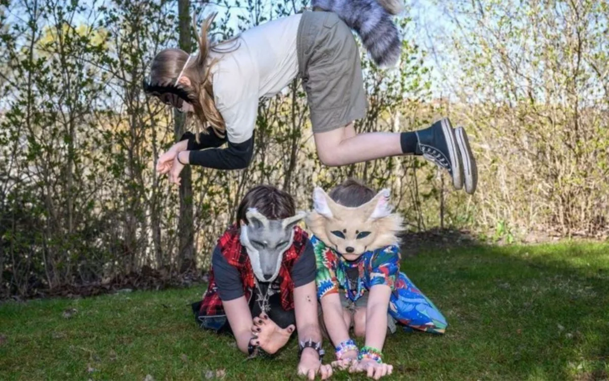 Tres jóvenes therians posan al aire libre en una zona de césped y arbustos. En el suelo, dos personas llevan máscaras de animales: una de zorro rubio y otra de lobo gris, mientras están arrodilladas. Una tercera persona salta por encima de ellas, congelada en el aire, vistiendo ropa informal y una gran cola de mapache peluda.